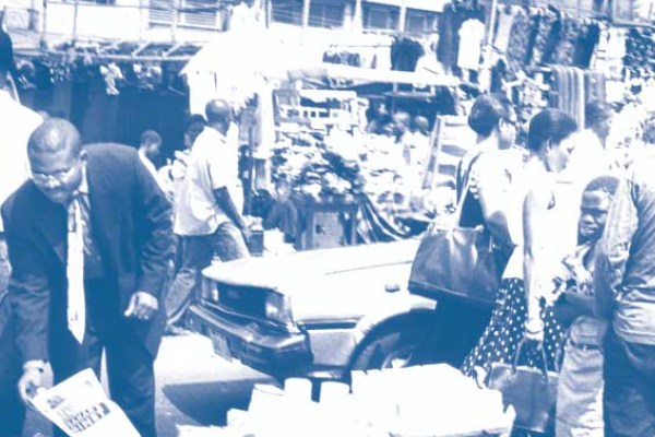 A duotone blue and white photograph of a bustling outdoor market. A man in a suit bends over in the foreground, while other people, a car, and market stalls are in the busy background.