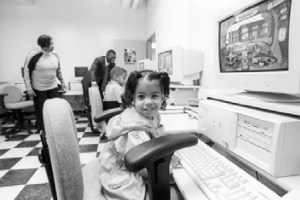 A black and white photo shows a young child with pigtails, sitting at a computer and smiling at the camera. In the background, two adults and another child are visible in a room with a checkered floor.