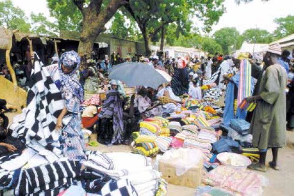 A vibrant outdoor marketplace with many people. Merchants sit behind piles of colorful, patterned textiles and other goods. One person holds a large umbrella for shade, and trees are visible in the background.