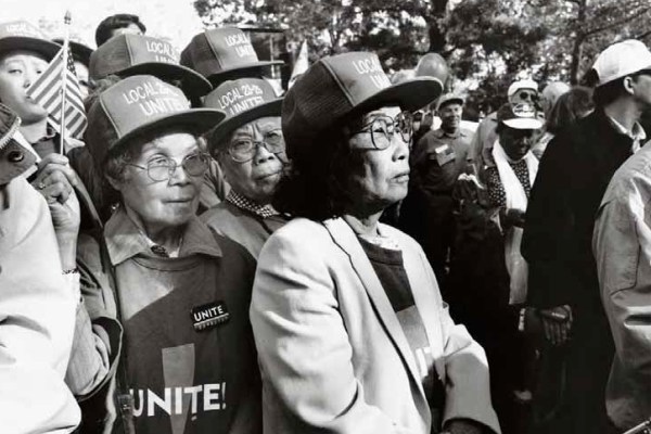 A black and white photo of a group of Asian American women at a rally or protest. They are wearing hats that say "LOCAL 23-25" and shirts with "UNITE" on them. They appear focused and determined.