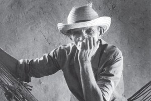 A black and white portrait of a man in a straw hat. He is leaning on a hammock, looking directly at the camera with one hand near his mouth. The background is a plain, textured wall.