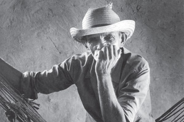 A black and white portrait of a man in a straw hat. He is leaning on a hammock, looking directly at the camera with one hand near his mouth. The background is a plain, textured wall.