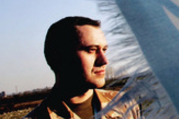 A portrait of a man with short hair, wearing a patterned shirt, looking to the right. A blurred, out-of-focus object is in the foreground, and the background shows an outdoor setting under a blue sky.