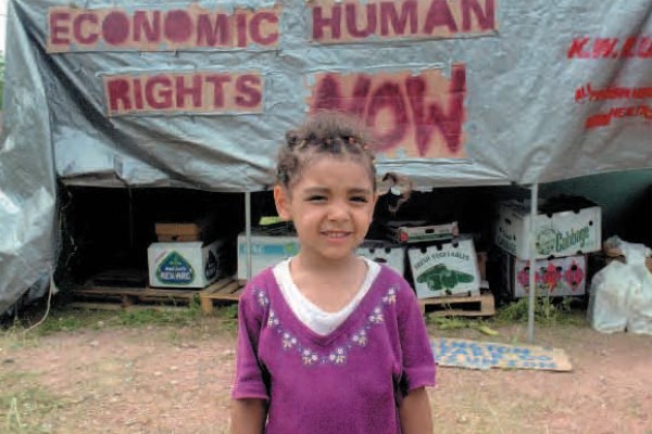 A smiling young girl with braided hair and a purple shirt stands in front of a gray tarp shelter. Behind her, cardboard boxes are stacked, and a banner reads "ECONOMIC HUMAN RIGHTS NOW."