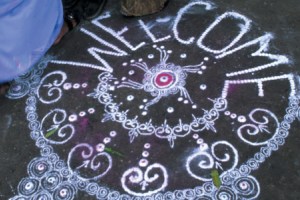 A chalk drawing, or rangoli, on a dark surface shows a circular floral pattern with the word "WELCOME" above it. The design is primarily white with small touches of pink and purple.