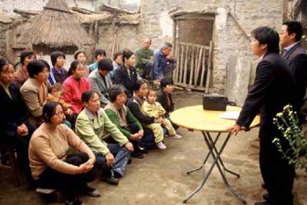 A group of about two dozen people, including women, children, and a few men, are gathered outdoors on a dirt ground, listening to two men in suits standing behind a small table.