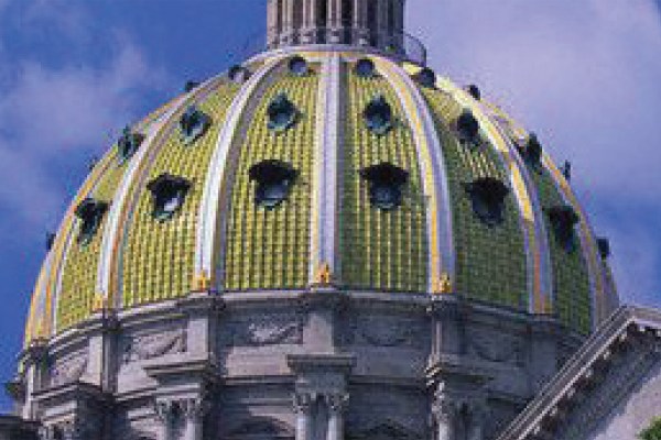 A close-up of a large, ornate dome, featuring a golden, tiled pattern and circular windows. The building below is made of gray stone, and the sky is a vibrant blue with some clouds.