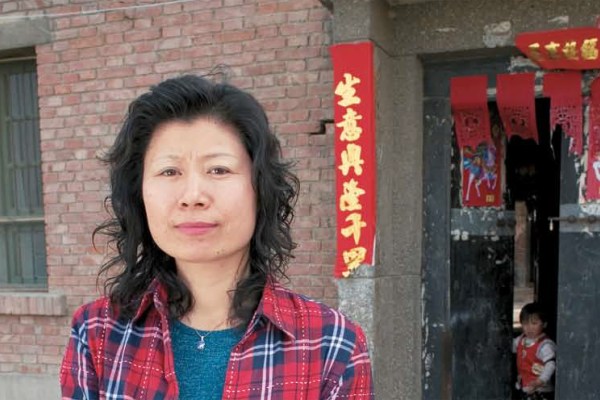 A head-and-shoulders portrait of a woman with dark hair, wearing a red plaid shirt, standing in front of a brick building. Red banners with Chinese characters hang beside the doorway, where a child is visible.