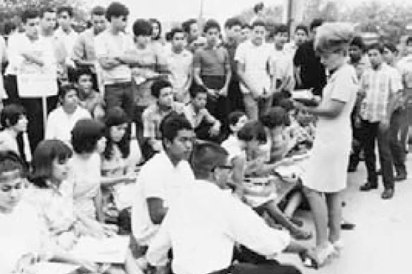 A black and white photo shows a large group of young people, many of whom are seated on the ground. A woman in a light-colored dress stands at the front, appearing to address the crowd.