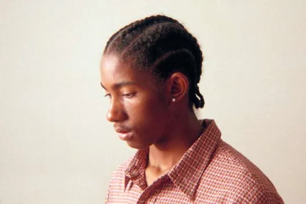 A head-and-shoulders portrait of a young person with braided hair, wearing a reddish-brown plaid collared shirt, looking down and to the left against a light, plain background.