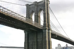 A photograph of the Brooklyn Bridge from a low angle, showing the towering stone arches and a network of suspension cables. The sky is slightly overcast, and a glimpse of the city is visible below.
