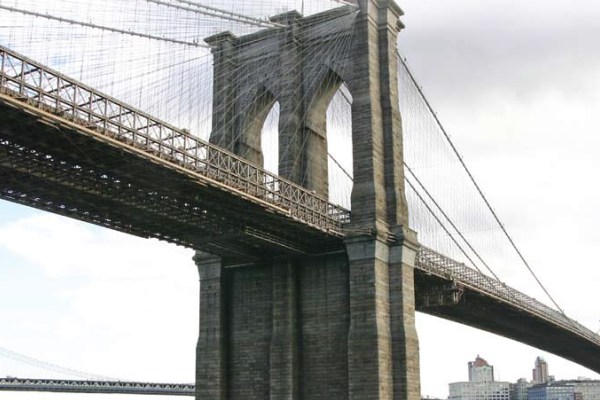 A photograph of the Brooklyn Bridge from a low angle, showing the towering stone arches and a network of suspension cables. The sky is slightly overcast, and a glimpse of the city is visible below.