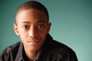 A portrait of a young person with short hair, wearing a denim jacket, looking directly at the camera. The background is a solid turquoise color.