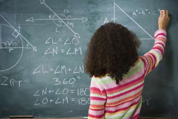 A student with curly dark hair and a colorful striped sweater stands at a blackboard, writing mathematical equations and drawing geometric shapes with chalk.