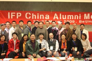 A group of about twenty people, most of whom are smiling, are seated and standing behind a long table. A large red banner behind them reads "IFP China Alumni Meeting" and "2009, Beijing."