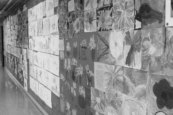 A black and white image of a long school hallway wall covered in various children's artworks. The pieces include paintings, drawings, and writings, with a collection of floral-themed art prominent in the foreground.
