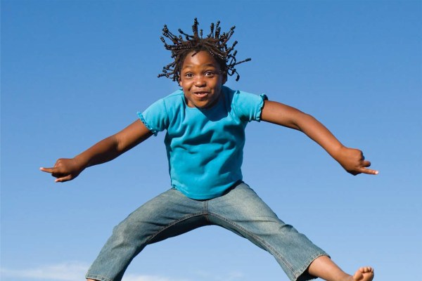 A child with braided hair is captured mid-air against a clear blue sky, smiling with a playful expression. They wear a blue t-shirt and jeans, with their arms and legs spread out.
