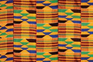 A colorful, tightly-woven textile with repeating vertical bands of geometric patterns. The colors include various shades of yellow, green, blue, brown, and red.
