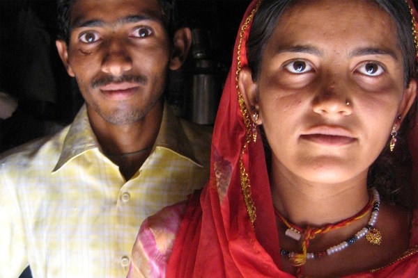 A close-up portrait of two people. A man in a yellow checkered shirt is slightly out of focus behind a woman in a red veil and jewelry, who looks directly at the camera.