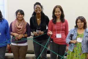 A diverse group of five women stands in a line, holding a green string in a complex web. They are all smiling or laughing, participating in a group activity in a well-lit indoor setting.