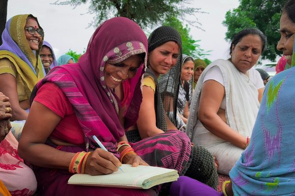 A group of Indian women in traditional colorful sarees seated outdoors together, with one smiling woman writing in a notebook during a community literacy or economic development gathering.