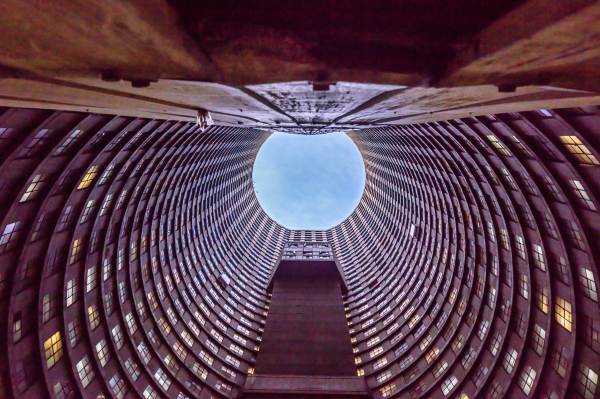 A worm's-eye view looking up through the hollow, circular core of the Ponte City Apartments in Johannesburg, South Africa. The towering brutalist architecture features rows of lit windows spiraling toward a bright blue sky, showcasing iconic urban design and South African landmarks.