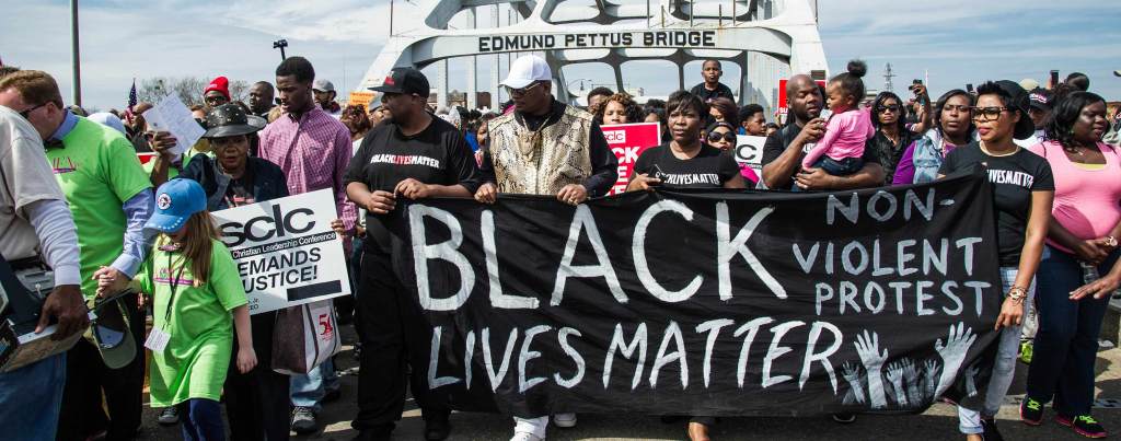 A large group of protestors walk together on Edmund Pettus Bridge. Multiple Black demonstrators hold a banner in front of them that reads "Black Lives Matter" and "Non-violent Protest". Another protestor holds a small cardboard sign that reads "Southern Christian Leadership Conference demands justice!"