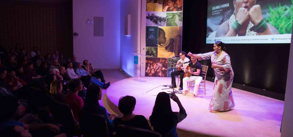 Hina Wong-Kalu dances hula on stage accompanied by Keali'i Reichel and Shawn Pimental playing guitar. Wong-Kalu wears a long gray dress with a maroon pattern. Behind the performers, a projecgtor screen shows a still of the documentary film, Kumu Hina.