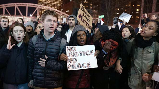 A crowd of people in winter coats protest at an Eric Garner demonstration. Two protesters hold signs that say "No Justice, No Peace" and "Black Lives Matter."