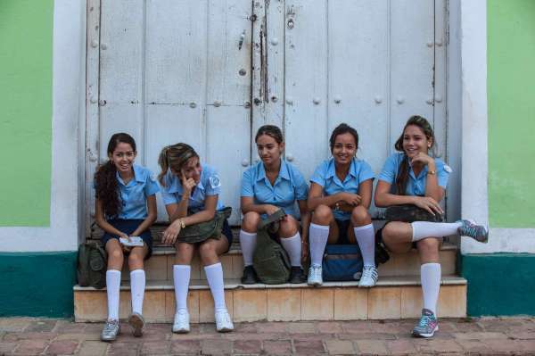 Five students with medium skin tones sit on a stoop in front of a close white gate. They wear identitical school uniforms with pale blue collared short-sleeved shirts and shorts with white socks and their hair pulled back into ponytails. 