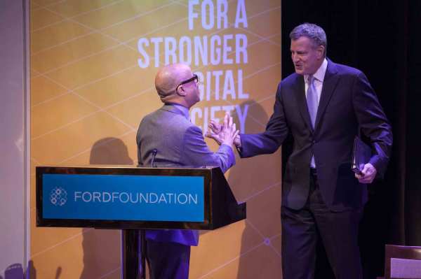 Bill deBlasio shakes Darren Walke's hand on-stage behind a podium with a Ford Foundation logo placard. A large banner that reads "For A Stronger Digital Society" hangs behind them.