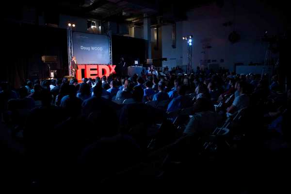 Doug Wood speaks onstage with a TEDx event. A large red TEDx logo is assembled behind him. He wears a dark colored suit and glasses.