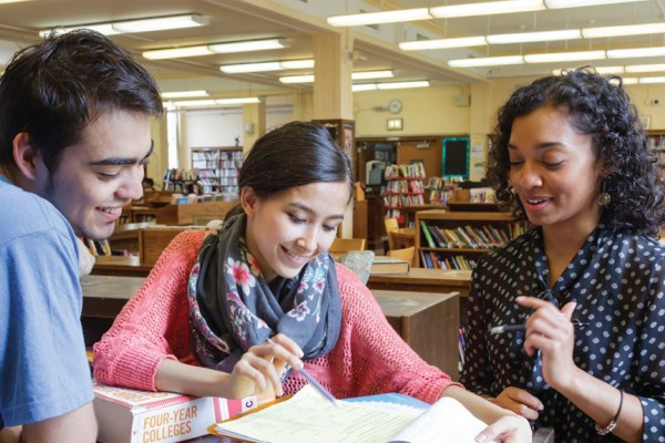 Three college-age students are seated at a table in a library, looking at a book on "FOUR-YEAR COLLEGES." The student in the center points to something in the book, while the other two look on and smile.