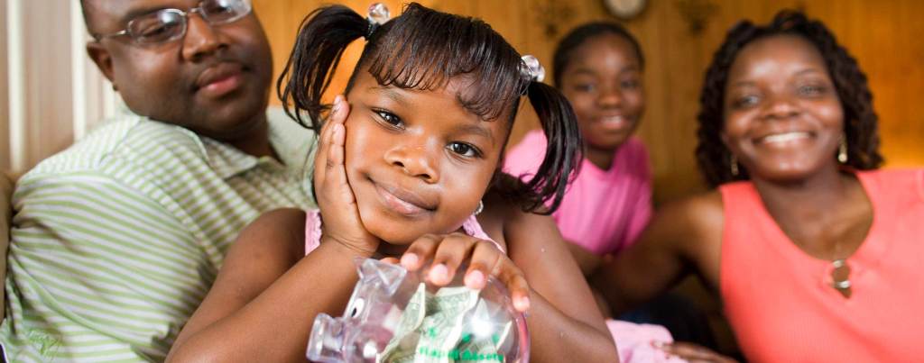 A young Black child in a pink shirt and pigtails poses with a clear piggy bank with money in it. Their family poses behind them, including a person with a mustache, eyeglasses, and a green striped shirt; another child in a hot pink t-shirt, and a smiling person with their hair in a bob, a coral pink tank top, and gold earrings.