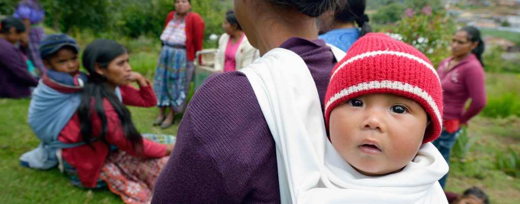 A small Maya Mam baby wrapped in a cloth sling on their parent's back and wearing a red and white knit beanie stares at the camera. The parent is gathered with other parents and children outdoors in a grass field.