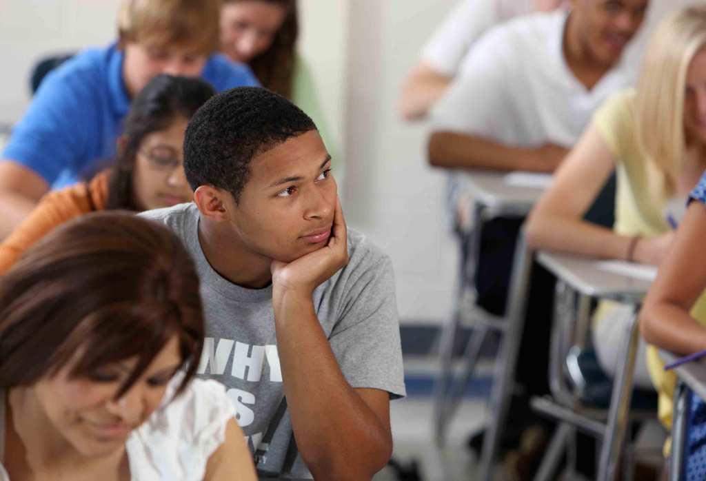 College students sit in desks in a classroom, focused on work. One student is looking up, elbow on the desk with their chin in their hand.