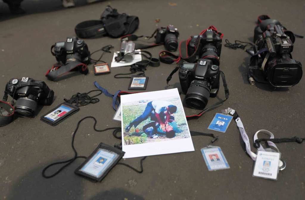 Black cameras and press credential badges are arranged on the floor around a photo of an Indonesian person in police uniform choking another person lying on the ground.