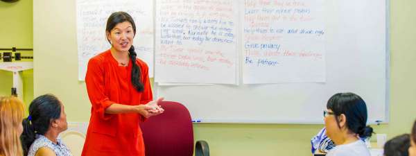 Ai-Jen Poo presents at the front of a classroom. There is a maroon swivel chair and whiteboard with large white note papers stuck to the wall that have writing on them. Ai-Jen wears a bright red dress and her black hair is a long braid. 