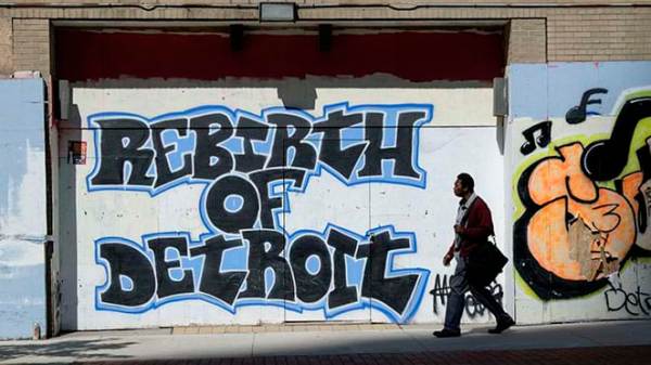 Grafitti that reads "Rebirth of Detroit" in bold black letters with a bright blue stroke decorates a wall. A person walks on the sidewalk past the street art.