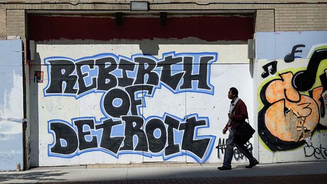 Grafitti that reads "Rebirth of Detroit" in bold black letters with a bright blue stroke decorates a wall. A person walks on the sidewalk past the street art.