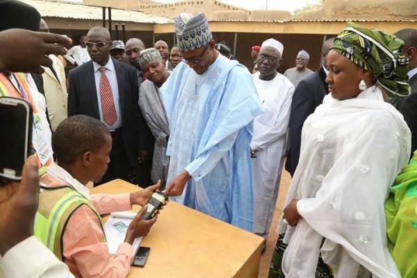 General Muhammadu Buhari dressed in light blue stands in front of a election worker, surrounded by other Nigerians. The crowd looks on as Buhari completes his accreditation for the governorship. 