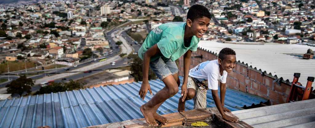 Two smiling children with medium dark skin tones and short hair play on the rooftop of a favela. They are barefoot, and wear t-shirts and shorts.