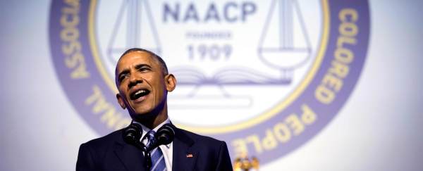 Barack Obama speaks into the mic. The seal of the NAACP is projected on the wall behind him. 