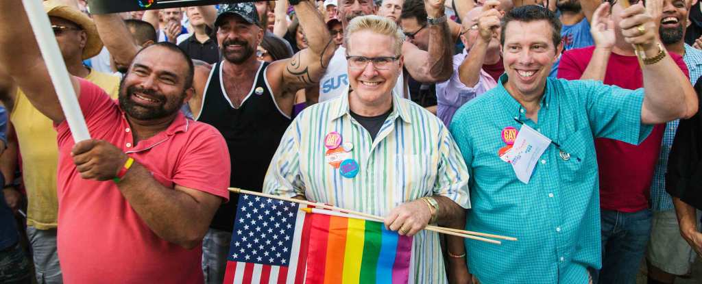 A crowd of smiling people in colorful clothing marches outside. A person in the middle wears a colorful striped short sleeve button down shirt with multiple LGBTQ+ pride buttons, and holds a U.S. flag and an LGBTQ+ pride rainbow flag.