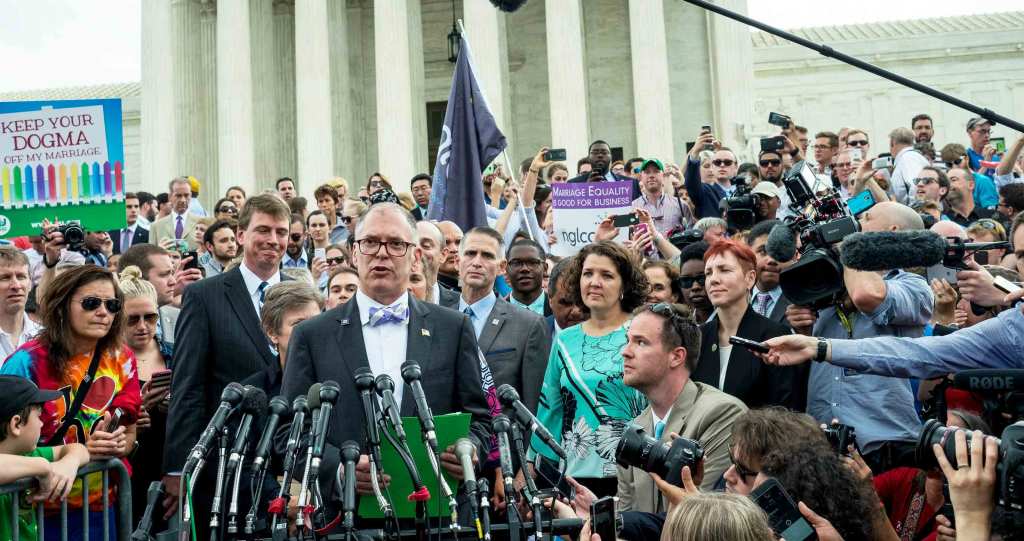 Jim Obergefell speaks to media in front of the Supreme Court. He is surrounded by activists and wears a suit and bowtie.