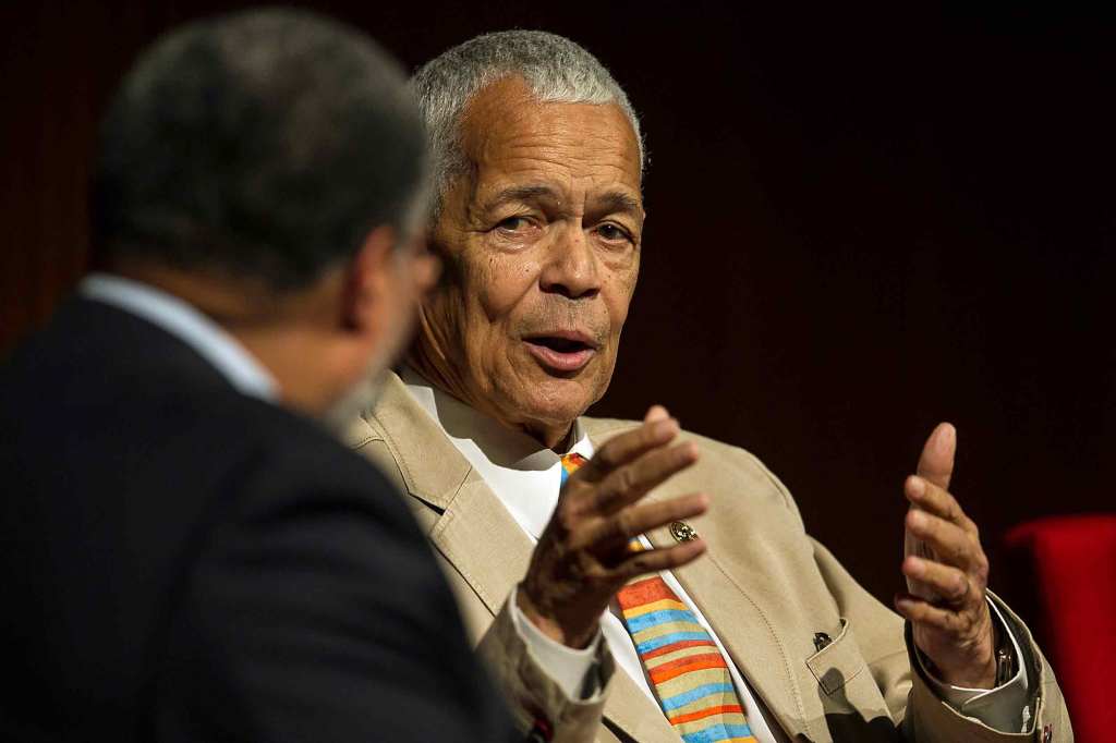 Julian Bond speaks to a moderator during an interview. He has short white hair and wears a khaki blazer and a colorful striped tie.