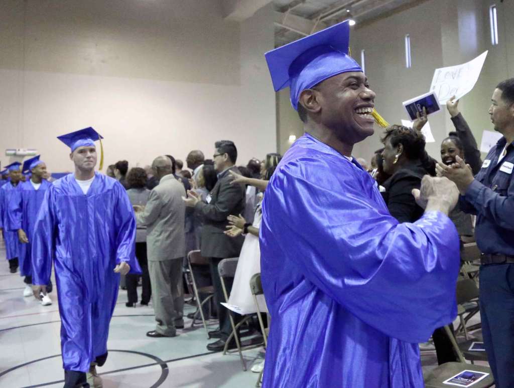 Happy graduates in blue caps and gowns with yellow tassels march down an aisle with smiles on their faces. An audience of people in folding chairs cheers for them.