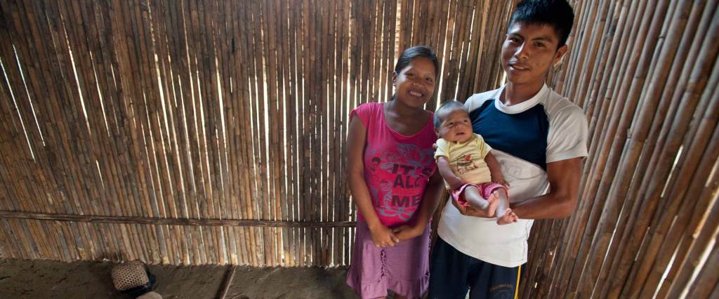 A young Peruvian couple smiles and poses with an infant baby.The mother wears a pink t-shirt and lavender skirt. The fater wears a white and blue t-shirt and matching pants. The baby he holds is wearing a yellow onesie and pinke shorts.