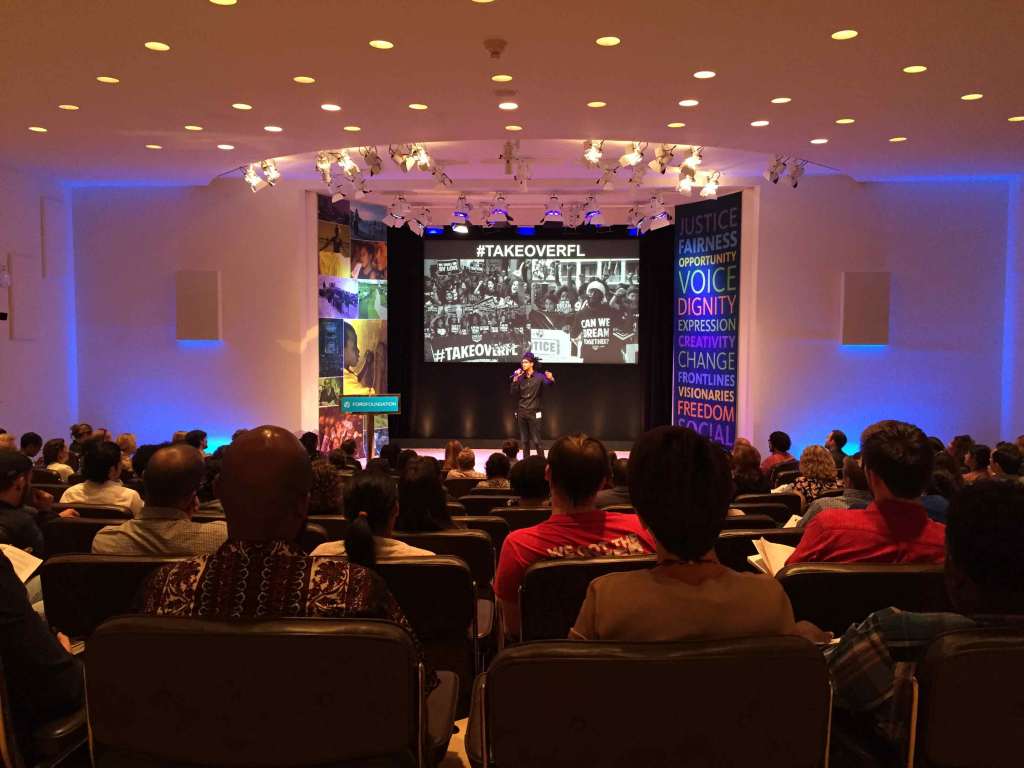 A person with medium dark skin tone stands on stage with a microphone, speaking to full audience in a theater. They stand in front of a projector screen with black and white photos of protesters  wearing shirts that say "Can we dream together?" with the hashtag #TakeOverFL.