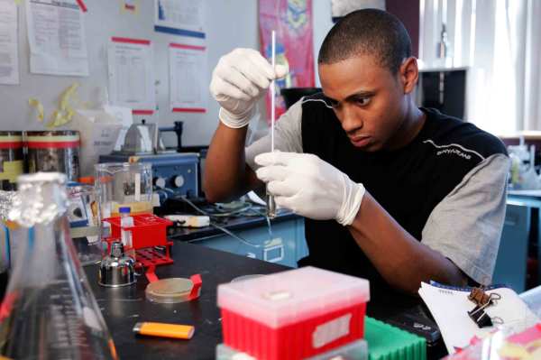 A Black student wearing white latex gloves holds a test tube and focuses intently on a pipette. They are in a chemistry lab surrounded by lab equipment. 